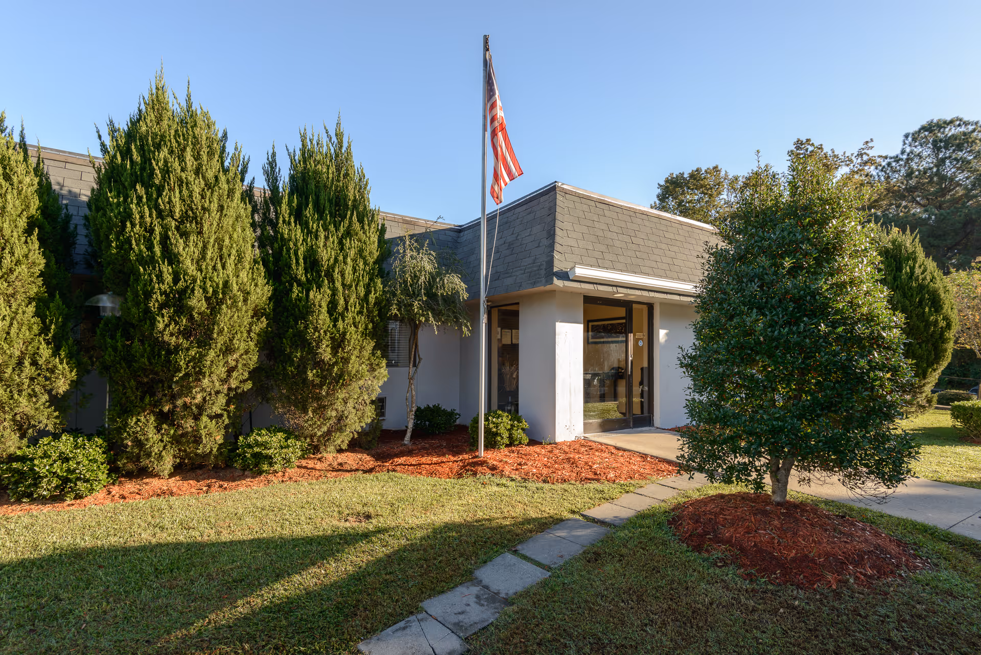 Front entrance of a low-rise building with a flagpole, glass entry door, and landscaped shrubs and lawn.