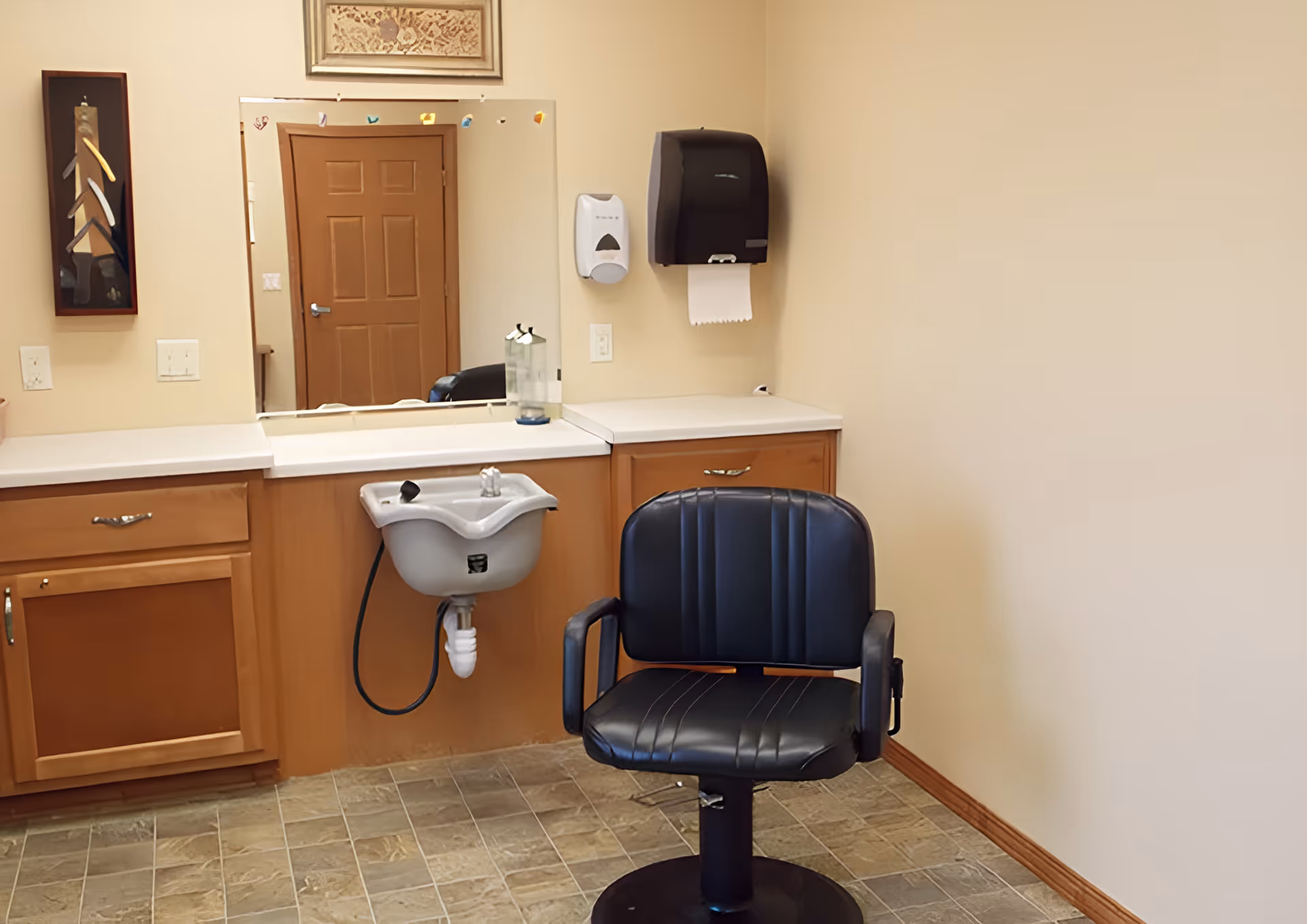 A small salon-style room with a black barber chair in front of a sink, mirror, countertop and wooden cabinets.