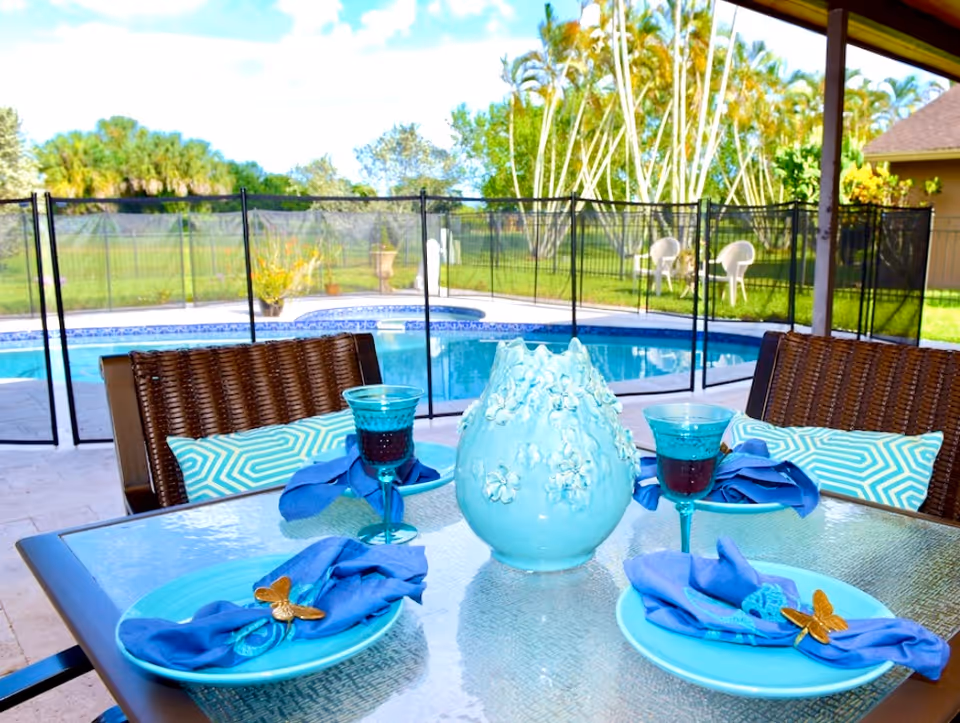 Outdoor patio dining area with a glass table set for three, featuring blue plates, blue napkins with butterfly napkin rings, and blue glasses. A decorative blue vase is in the center of the table. In the background, there is a fenced swimming pool, green lawn, and some white chairs near a small building under a partly cloudy sky.