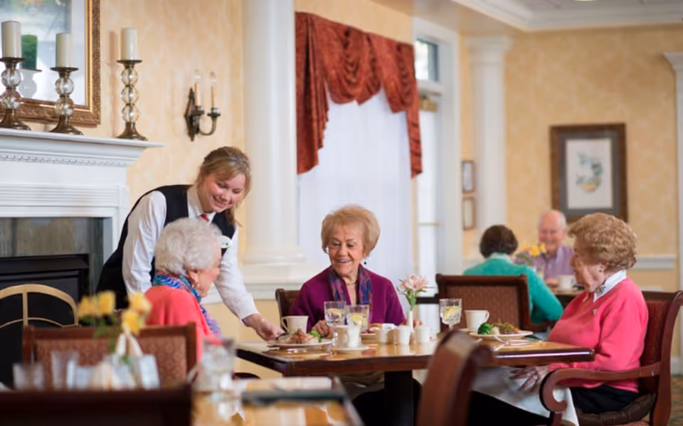 A dining room scene in a senior living facility where three elderly women are seated at a table enjoying a meal. A smiling waitress is serving food to one of the women. The room has warm yellow wallpaper, a white fireplace with candles, framed artwork, and red curtains on the windows.