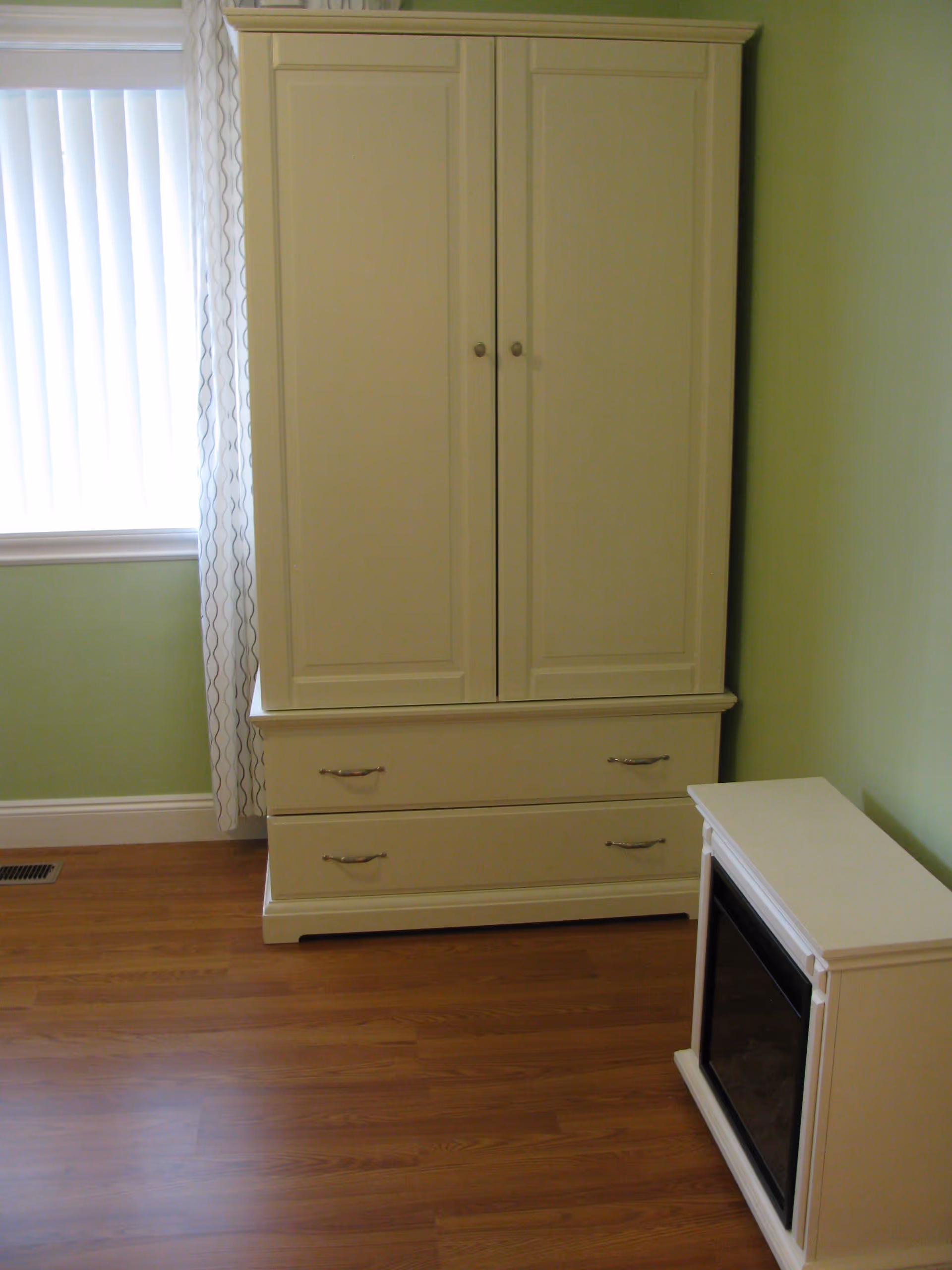 A corner of a room with light green walls and wood flooring. There is a white wardrobe with two doors and two drawers, next to a small white electric fireplace. A window with vertical blinds and a sheer curtain is partially visible on the left side.