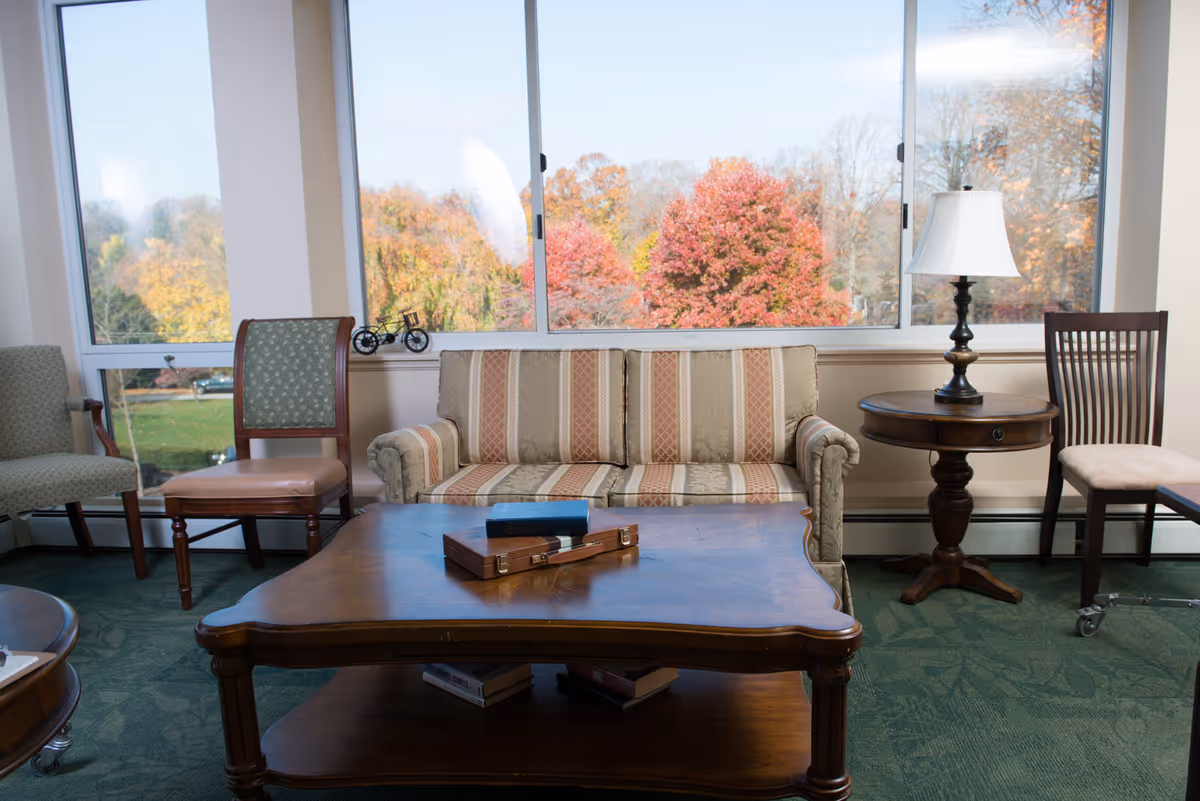 A cozy sitting area with a striped sofa, two wooden chairs, a round side table with a lamp, and a wooden coffee table with books and a decorative box. Large windows behind the sofa show colorful autumn trees outside.