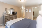 A bedroom with a neatly made bed featuring a dark blue blanket and white pillows. Across from the bed is a wooden dresser with a large mirror, a small lamp, and a flat-screen TV. The room has beige walls and carpeted floors. There are two open doorways visible, one leading to a closet and the other to another room or hallway. Two framed pictures hang on the wall between the doorways.
