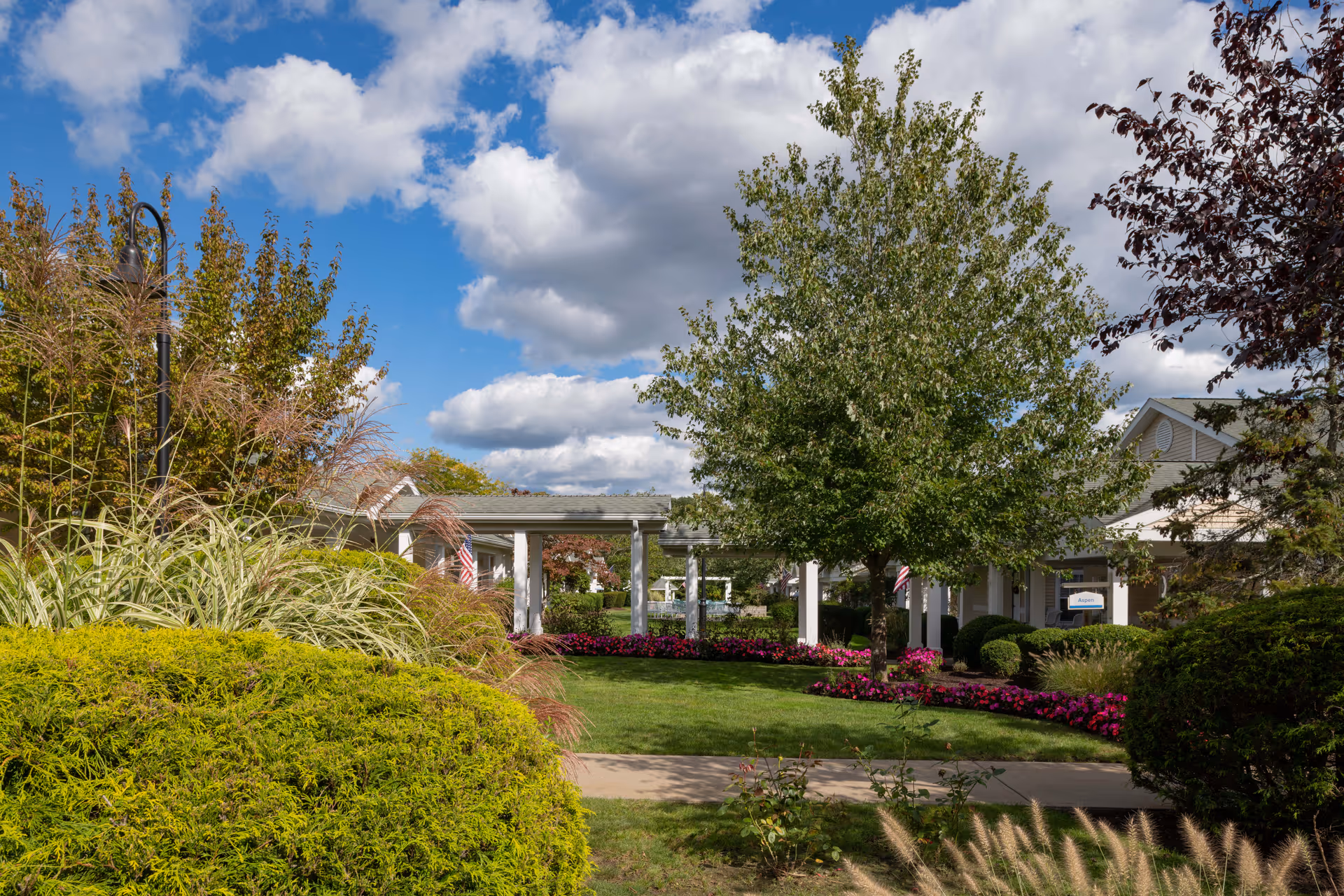 Front entrance of The Cottages of Dartmouth Village with a covered driveway, manicured lawns, trees and colorful flowerbeds under a partly cloudy sky.