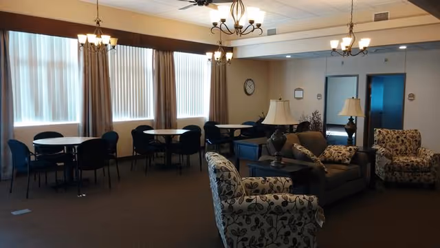 A spacious common area in an assisted living facility with several round tables and chairs near large windows with curtains. The room also features a seating area with a sofa, two patterned armchairs, side tables, and lamps. The walls are light-colored, and there is a ceiling fan and chandelier lighting fixtures.