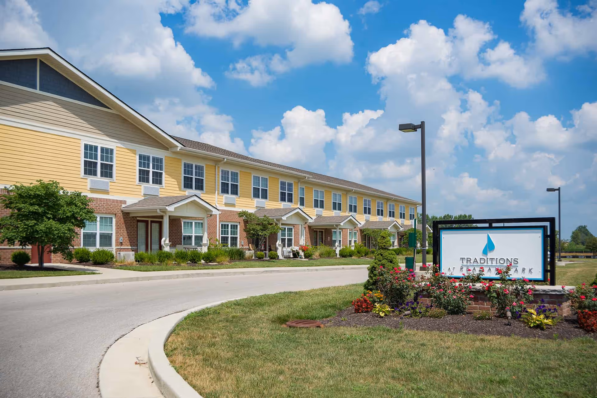 Front exterior of a two-story yellow and brick senior living building with landscaped flower beds and a 'Traditions at Reagan Park' sign.