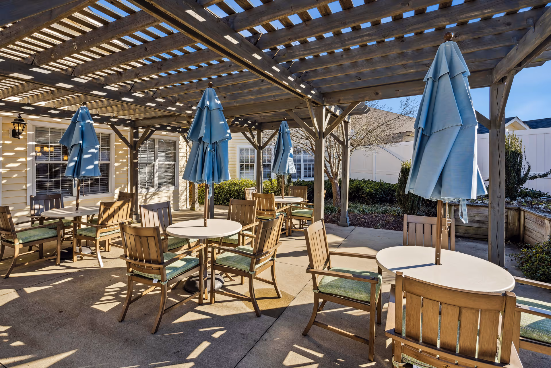 Outdoor patio area with round tables and wooden chairs under a wooden pergola. Each table has a closed blue umbrella. The patio is adjacent to a building with windows and surrounded by greenery and a white fence.