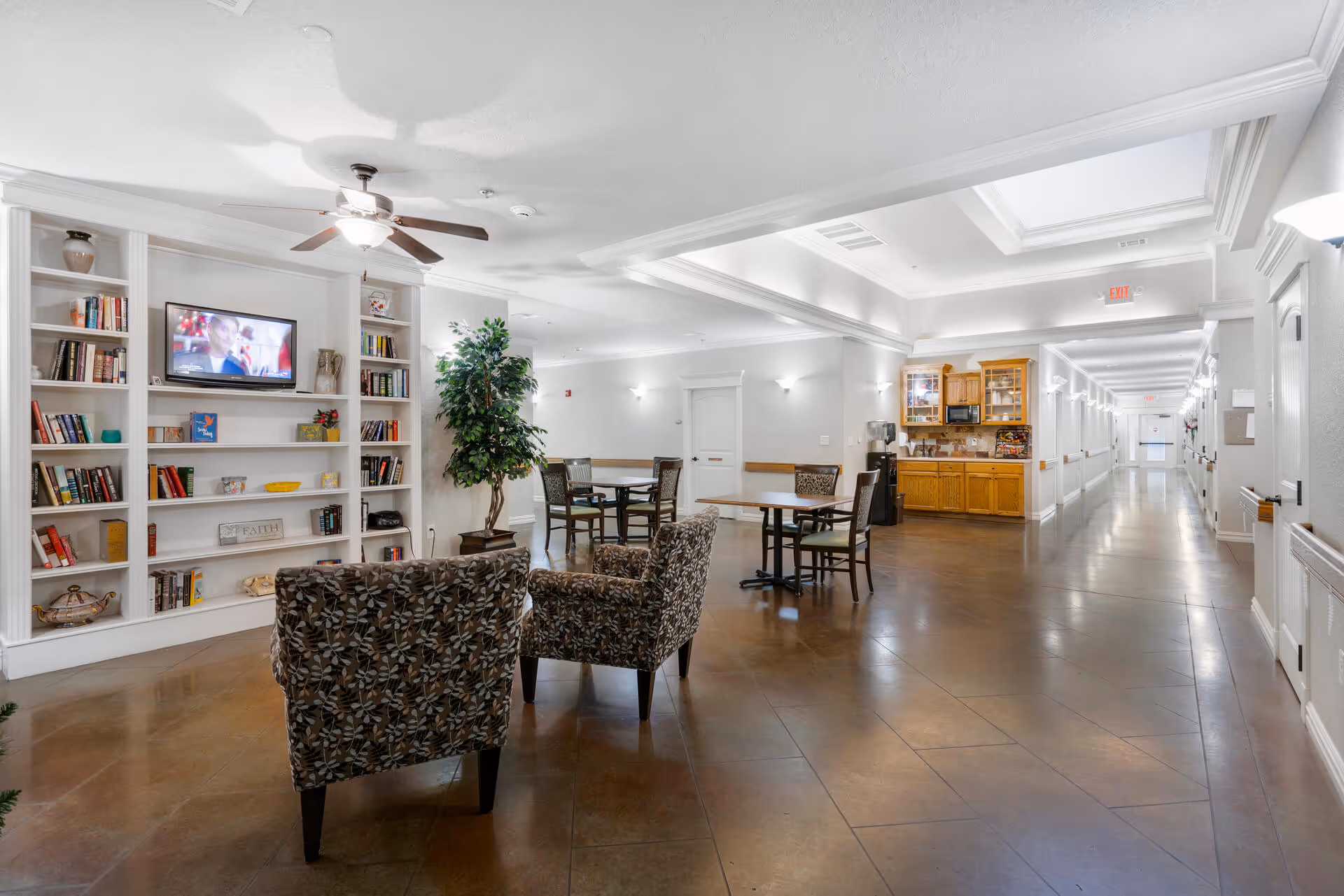 A bright communal lounge with bookshelves, patterned chairs, tables, a wall-mounted TV, and a kitchenette opening onto a long hallway.