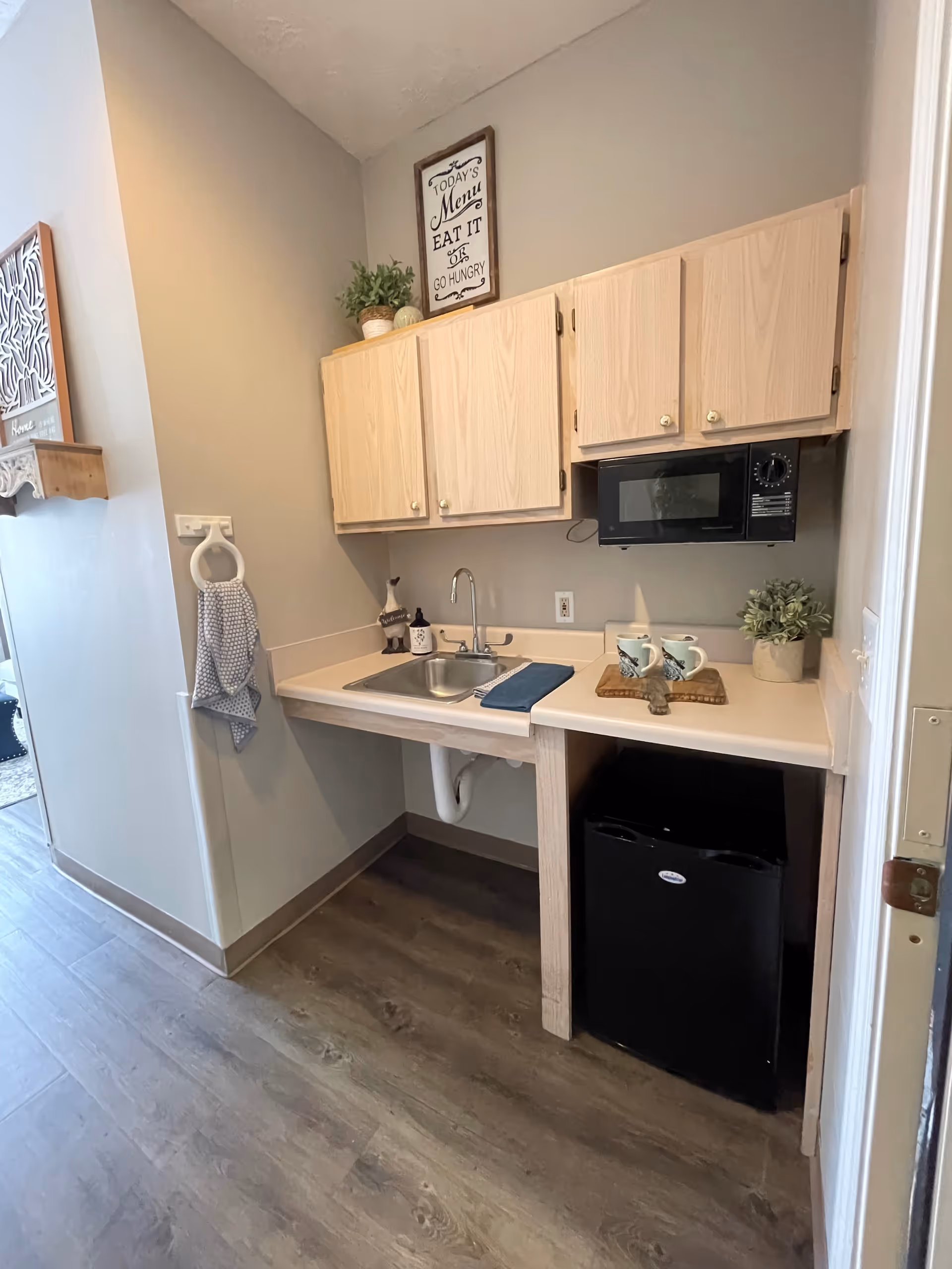 Small kitchenette area with light wood cabinets, a stainless steel sink, a black microwave mounted under the cabinets, a black mini refrigerator below the counter, two mugs on a wooden tray, a small potted plant, and a hand towel hanging on a ring on the wall. The floor is wood-style laminate and the walls are painted light gray.