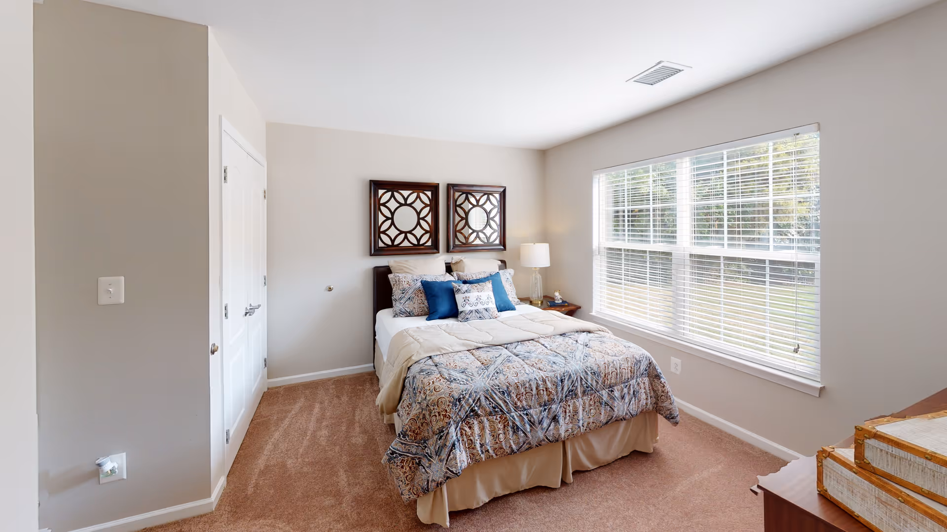 Bright bedroom with a made bed topped with decorative pillows, two framed wall mirrors above the headboard, a nightstand with a lamp, and a large window with blinds.