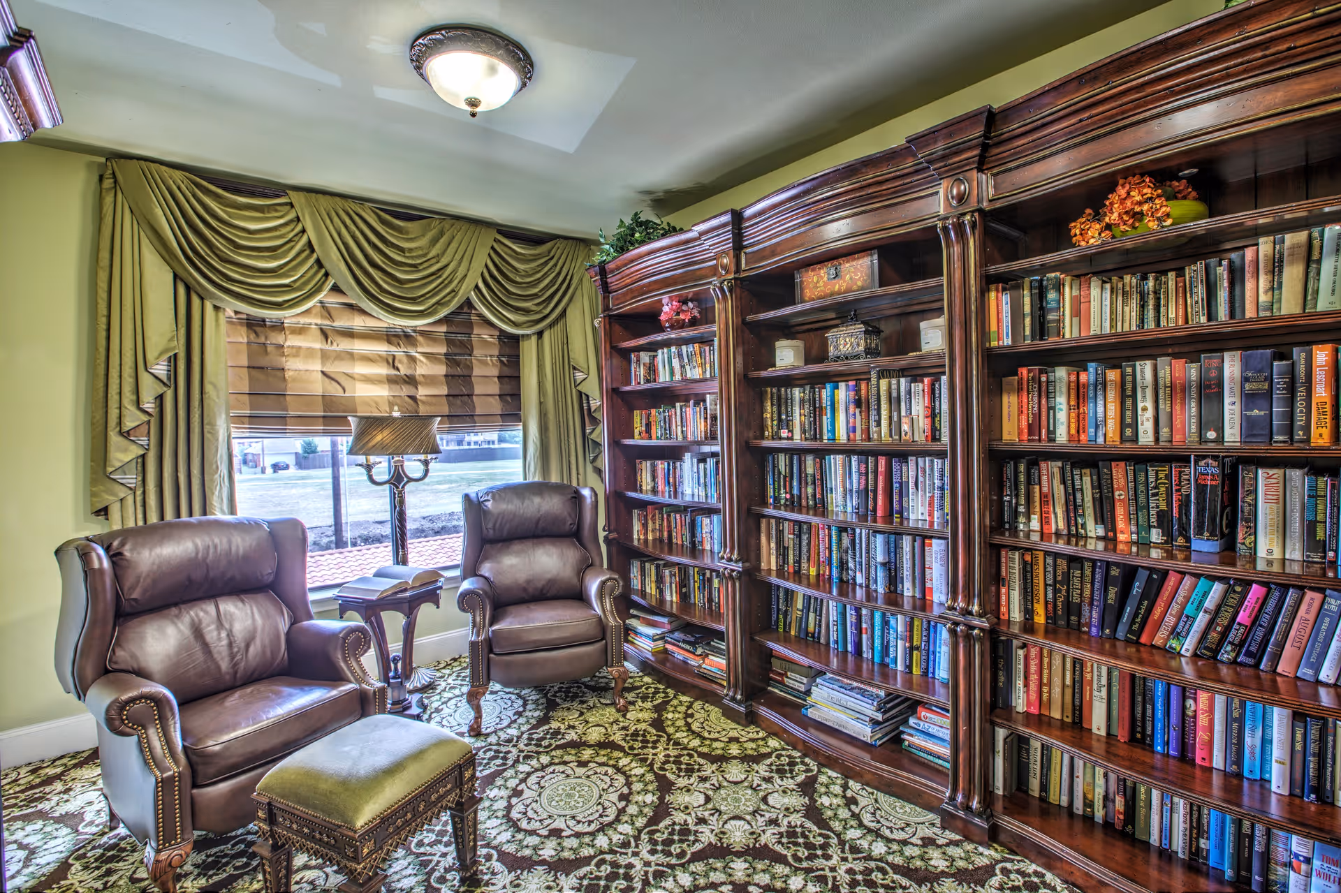 A cozy reading room with two brown leather armchairs and a matching ottoman placed on a patterned carpet. Behind the chairs is a large window with green drapes and a Roman shade. To the right, there is a large wooden bookshelf filled with numerous books and some decorative items on top.