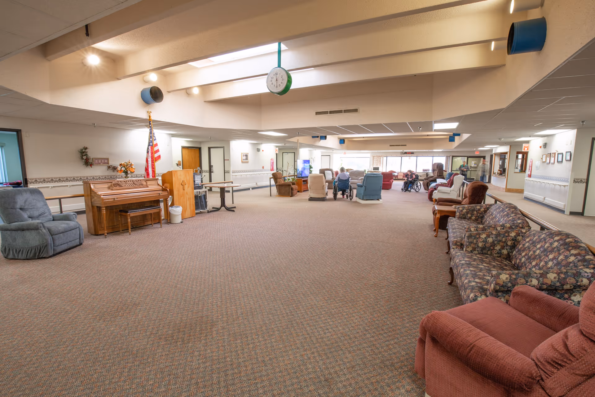 Spacious senior living common room with sofas and recliners, a piano, an American flag, and residents seated near the windows.