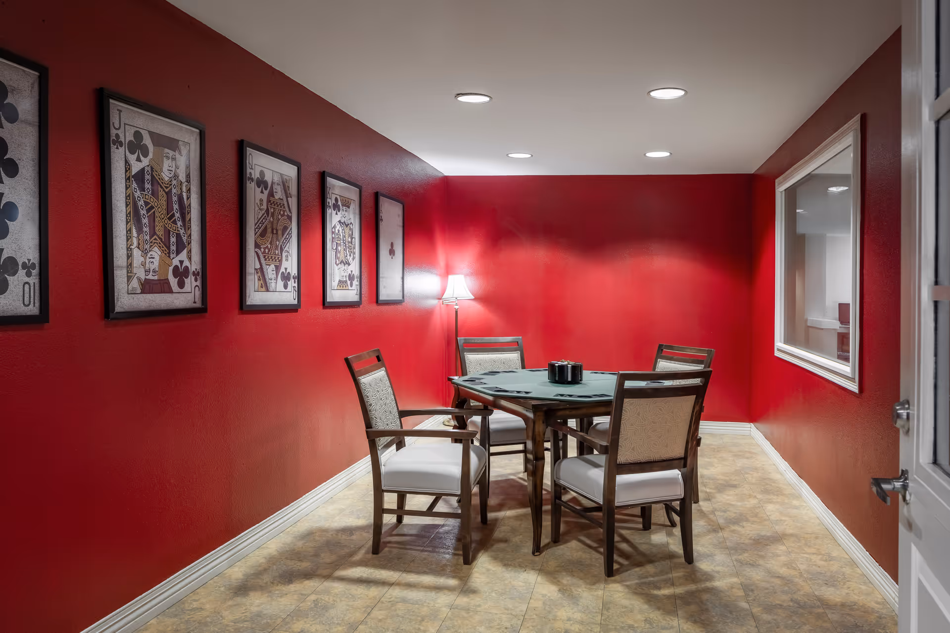 A small room with red walls featuring framed playing card artwork on one side. In the center, there is a round table with a green felt cover and four wooden chairs with cushioned seats. A floor lamp is placed in the corner, and there is a window on the right wall looking into another room.