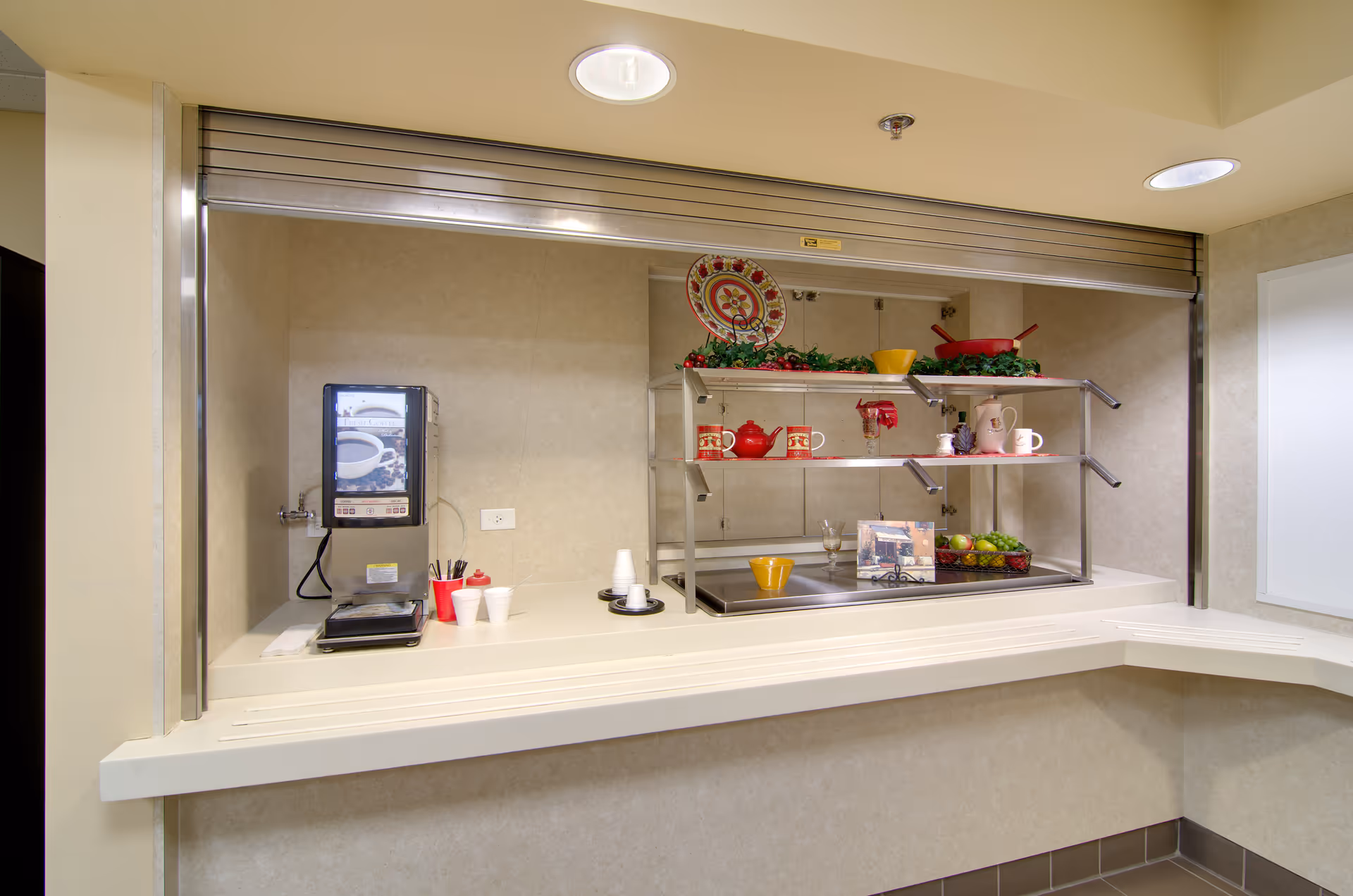 A food service pass-through counter with a coffee machine, shelving displaying dishes and decorative fruit, and a countertop service area.