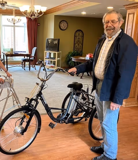 An elderly man standing indoors next to a black electric bicycle, smiling at the camera. The room has wooden flooring, a carpeted area with tables and chairs, a TV on a stand, and decorative wall elements. Another person with a walker is partially visible on the left side.