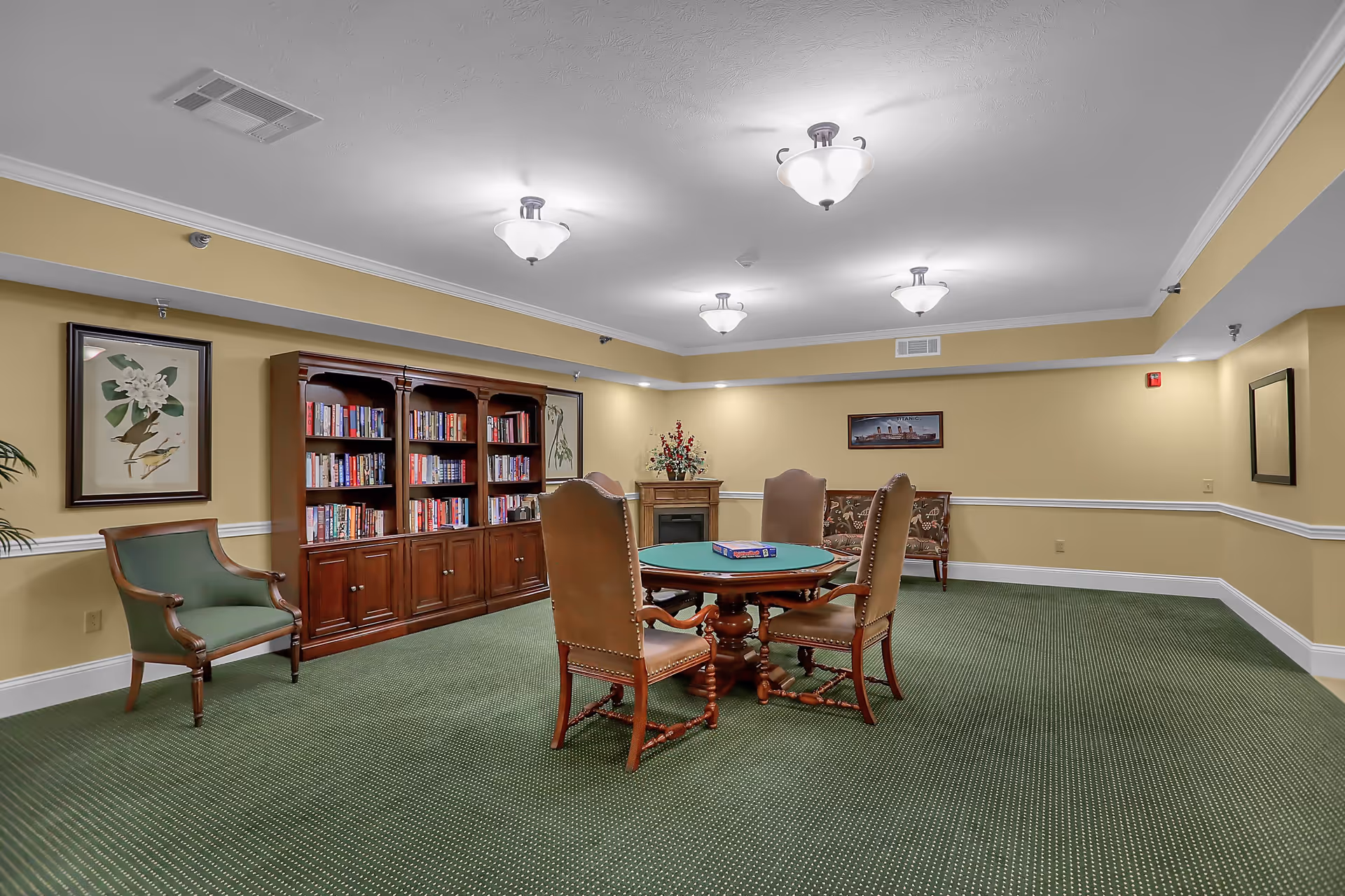A cozy interior room with green carpet and yellow walls featuring a wooden bookshelf filled with books, a round table with four chairs around it, a green armchair, a floral bench, framed artwork on the walls, and ceiling lights providing warm illumination.
