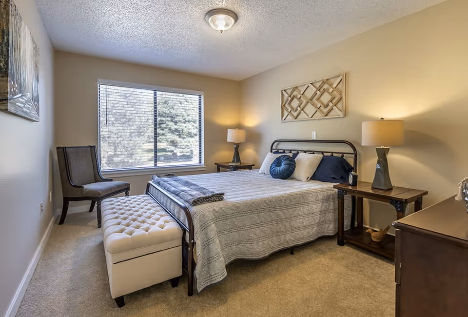 A cozy bedroom with a large window letting in natural light. The room features a bed with a metal frame, gray bedspread, and multiple pillows. There are two wooden nightstands with matching lamps on either side of the bed. A cushioned bench is placed at the foot of the bed, and a single upholstered chair is positioned near the window. The walls are painted beige and decorated with a framed artwork and a decorative wall piece above the bed.