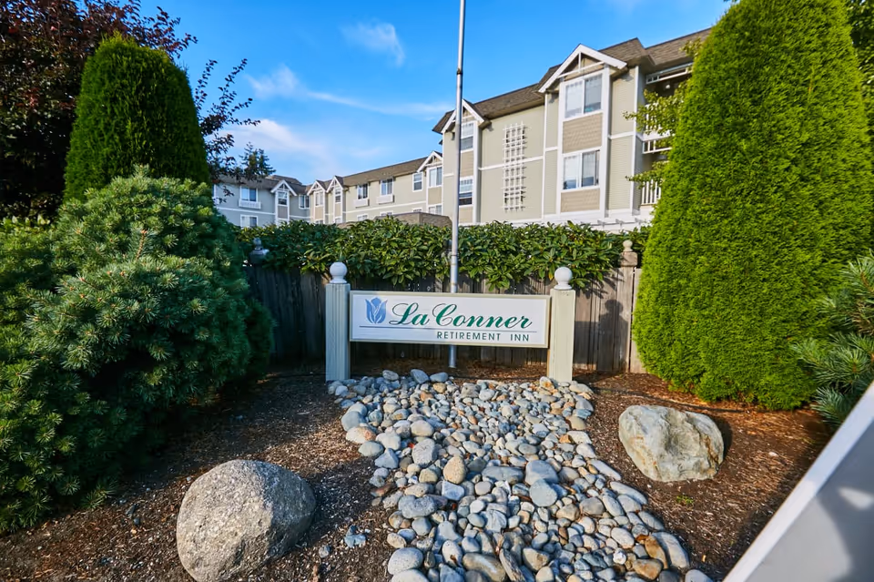 Outdoor view of La Conner Retirement Inn sign surrounded by landscaping with rocks, bushes, and trees, with the multi-story retirement building in the background under a blue sky.