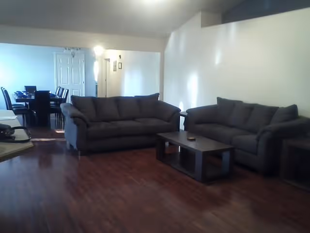 A living room with two dark gray sofas facing each other and a dark wooden coffee table in between. The room has wooden flooring and white walls. In the background, there is a dining area with a table and chairs, and a white door is visible.