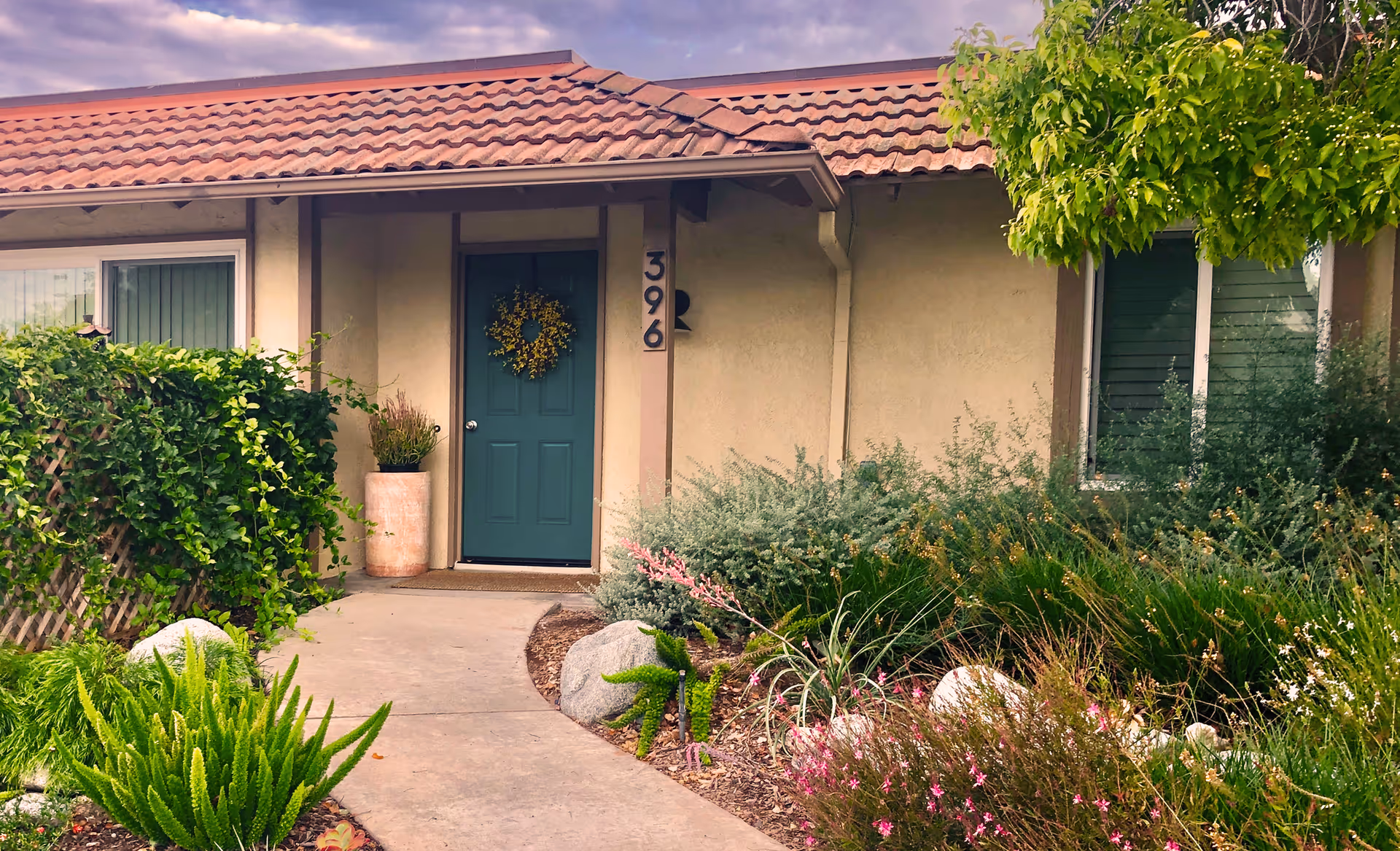 Front entrance of a single-story building with a green door decorated with a yellow wreath, surrounded by lush greenery and flowering plants. The building has a tiled roof and beige walls, with the number 396 displayed next to the door.