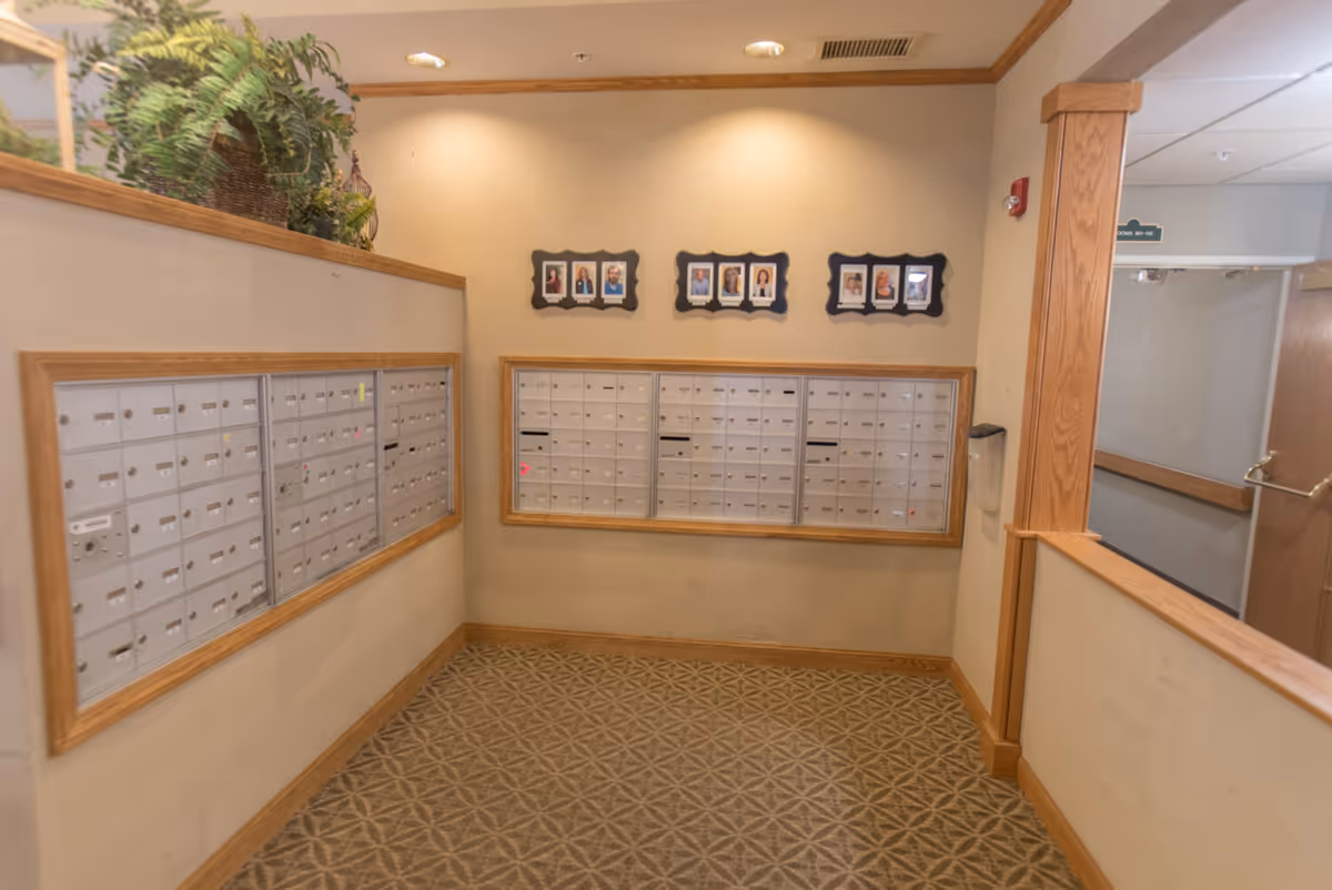 Interior view of a mailroom area with multiple mailboxes mounted on two walls. Above the mailboxes are framed photos of staff members. The room has beige walls, patterned carpet, and wooden trim. There is a plant on a ledge above one set of mailboxes and a hallway visible to the right.