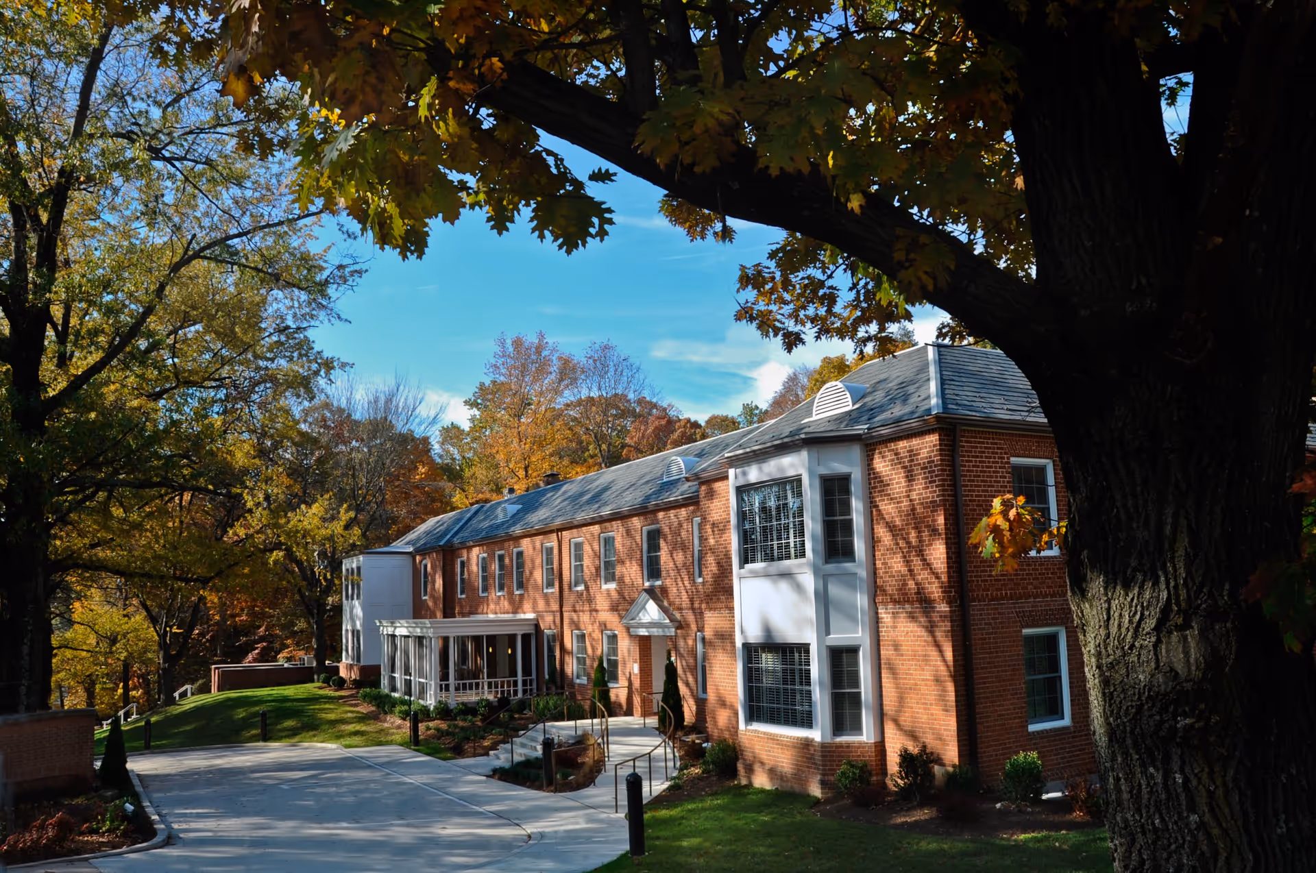 Two-story red brick building with a driveway, surrounded by large oak trees showing autumn foliage.