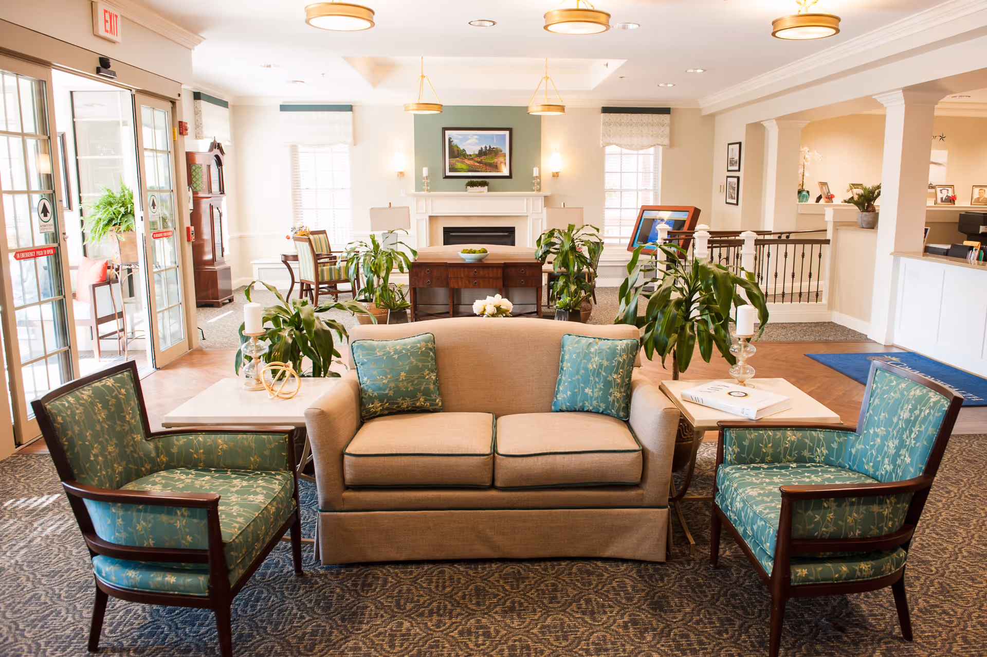 A bright and welcoming senior living facility common area with a beige loveseat and two green patterned armchairs arranged around two small white tables. The room features a fireplace with a framed landscape painting above it, several potted plants, and large windows letting in natural light. There is a reception desk visible to the right and glass doors to the left leading outside.