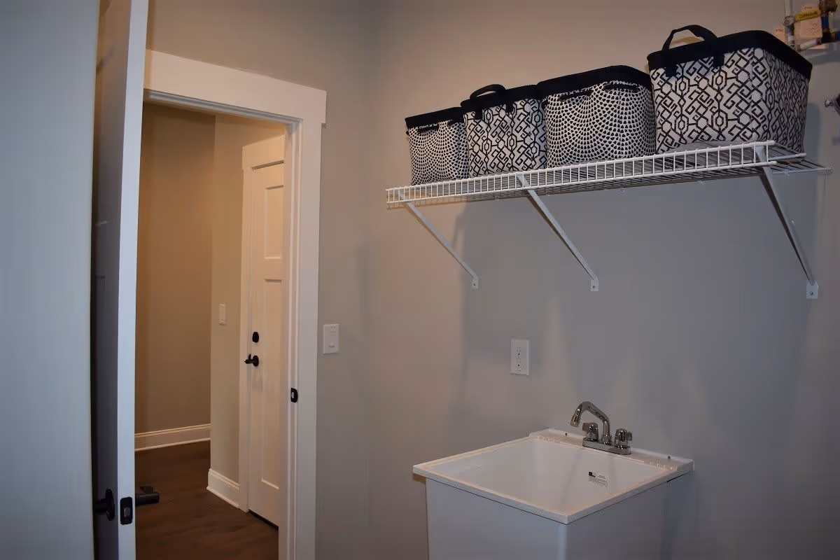 A laundry room with a white utility sink and a wall-mounted wire shelf above it holding four patterned storage baskets. The room has light gray walls and a doorway leading to a hallway with white doors and dark wood flooring.