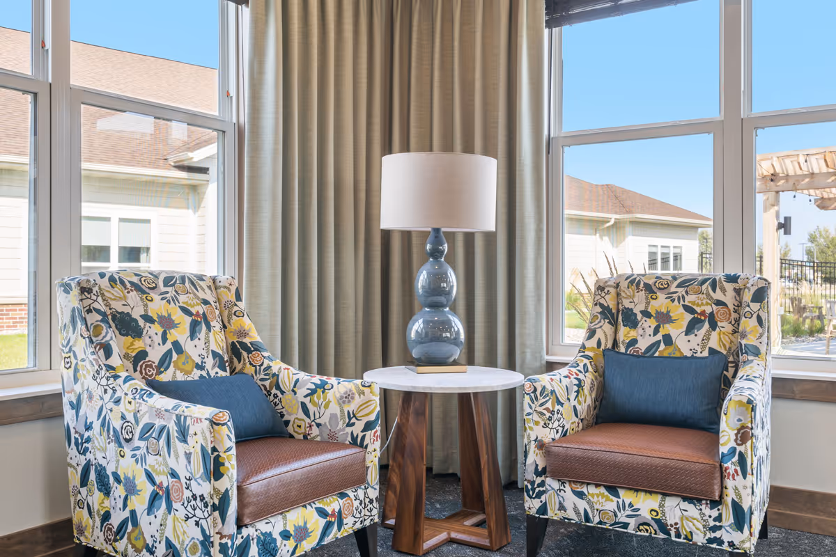 Two floral patterned armchairs with brown leather seats and blue cushions are positioned on either side of a round wooden table with a blue ceramic lamp on top. The seating area is in front of large windows with beige curtains, allowing natural light to fill the room and showing an outdoor view of buildings and a pergola.