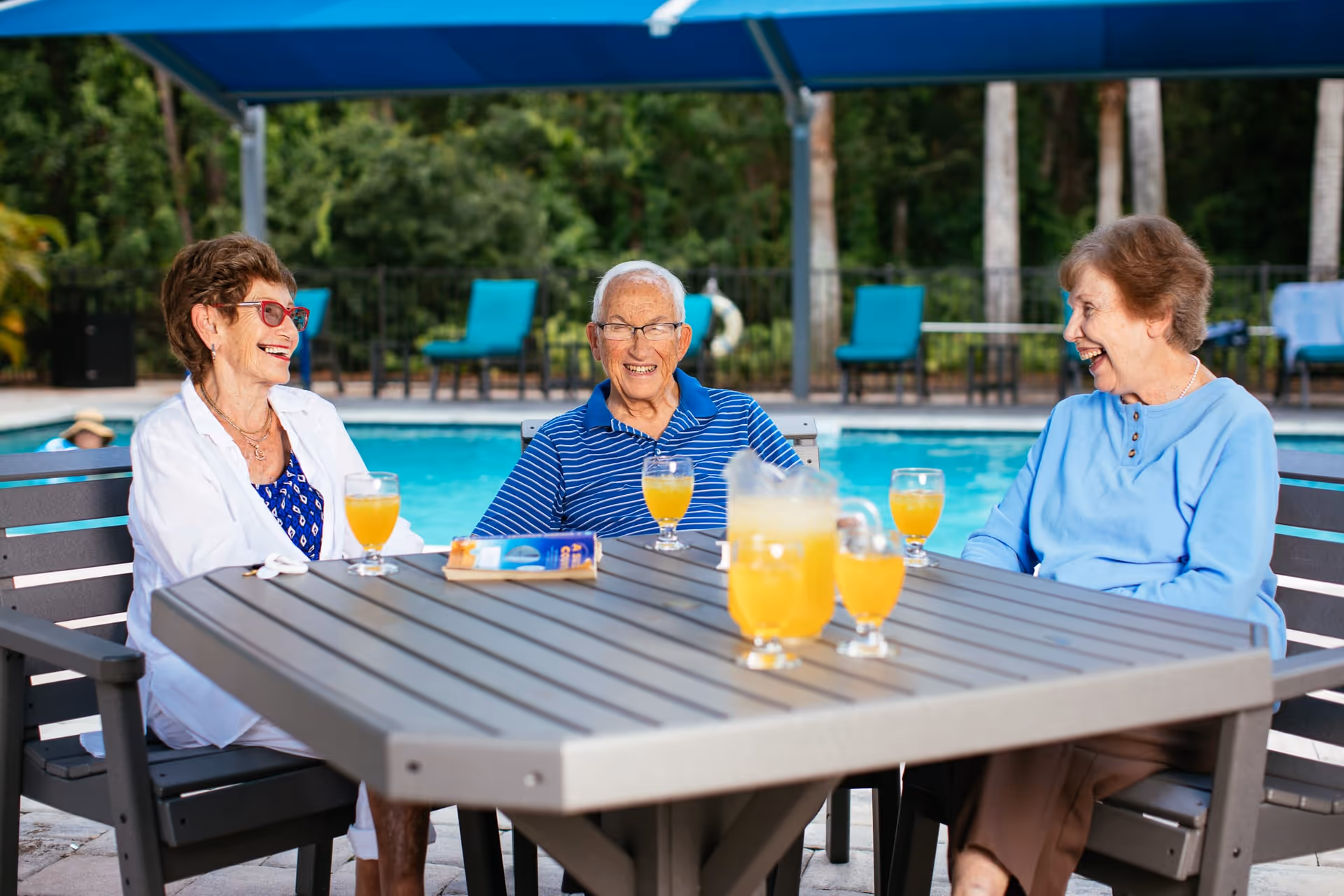 Three elderly people sitting around a table outdoors by a swimming pool, smiling and enjoying glasses of orange juice under a blue canopy with trees in the background.