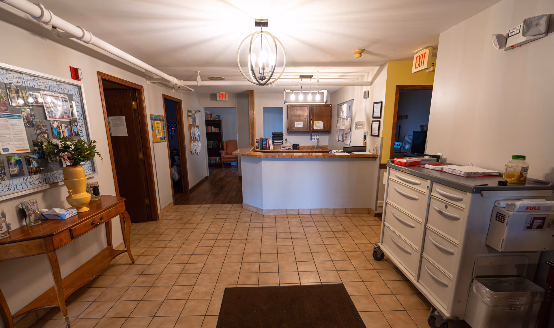Interior view of a reception area in an assisted living facility with a tiled floor, a wooden table with a yellow vase and flowers on the left, a reception desk straight ahead, and a white storage cart on the right. There are doors and hallways visible in the background along with exit signs and overhead lighting fixtures.