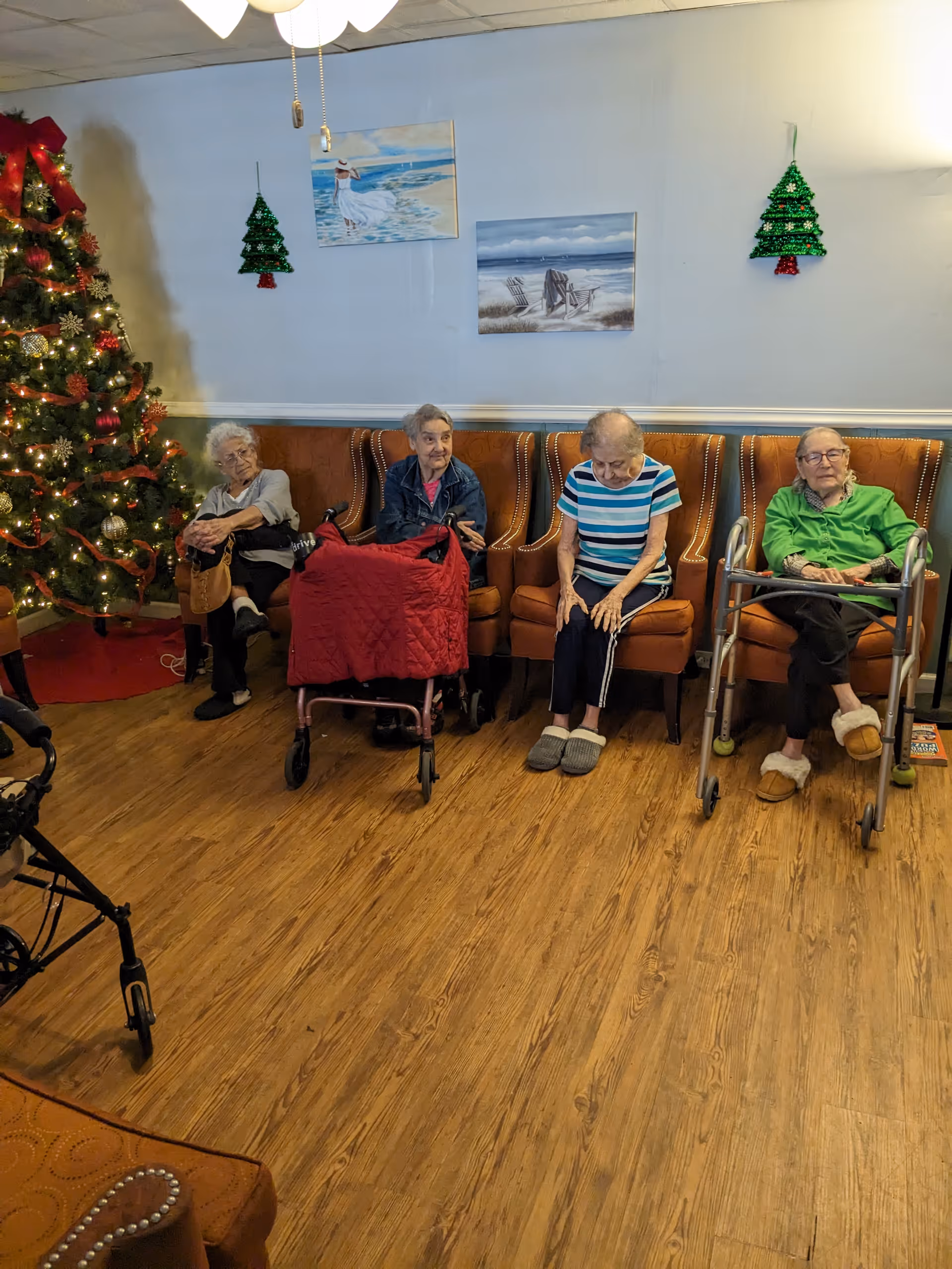 Four elderly women sitting in a row on brown armchairs in a room with wooden flooring. Two of the women have walkers in front of them. There is a decorated Christmas tree with lights and ornaments on the left side of the image. The wall behind them is light blue with two paintings of beach scenes and two green Christmas tree decorations hanging. The room has a cozy and festive atmosphere.