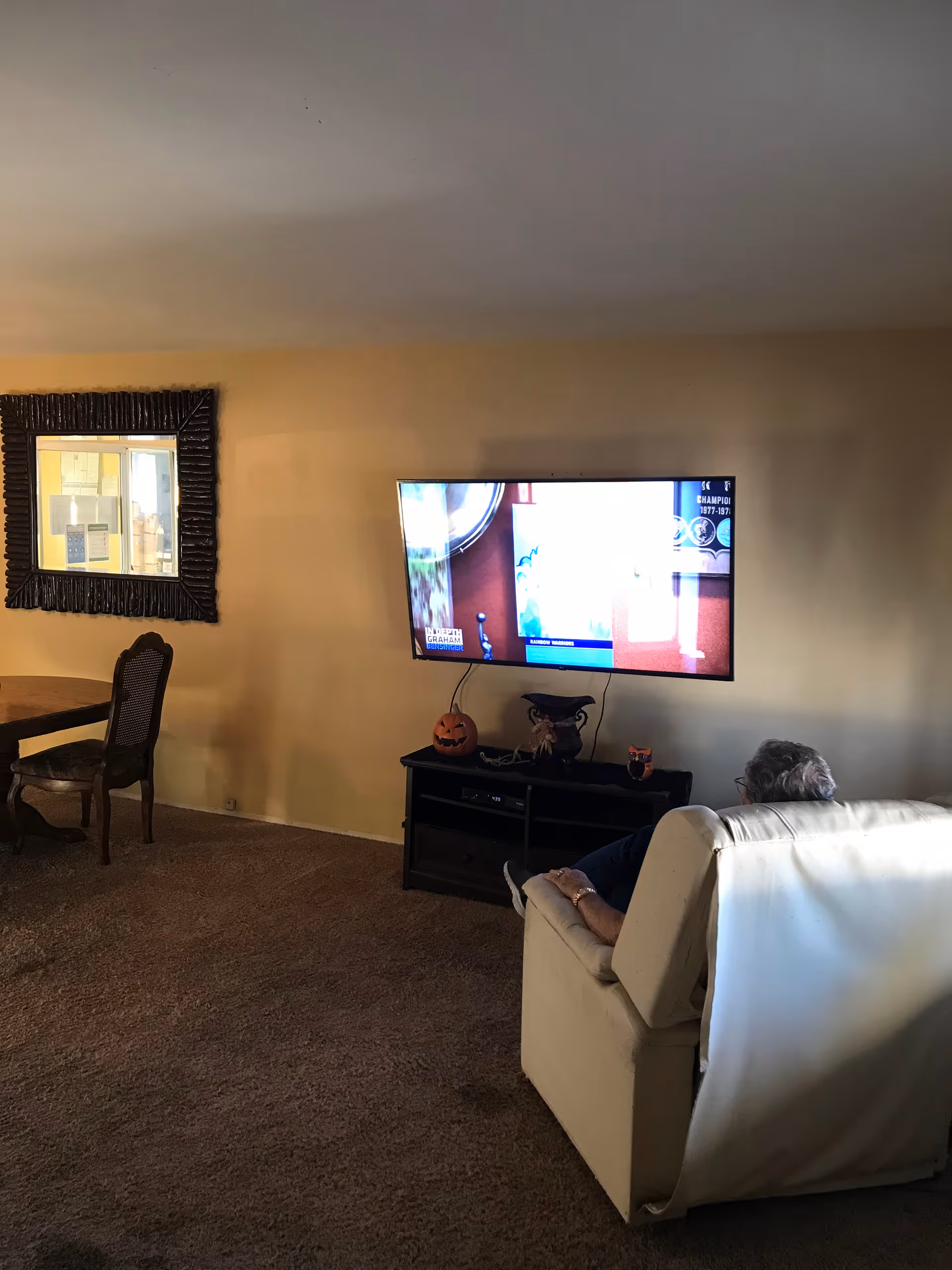 A person sitting in a white armchair watching a wall-mounted flat-screen TV in a living room. The room has beige walls and carpet, a wooden dining table with chairs, a large decorative mirror on the wall, and a TV stand with Halloween-themed decorations including a pumpkin and an owl figurine.