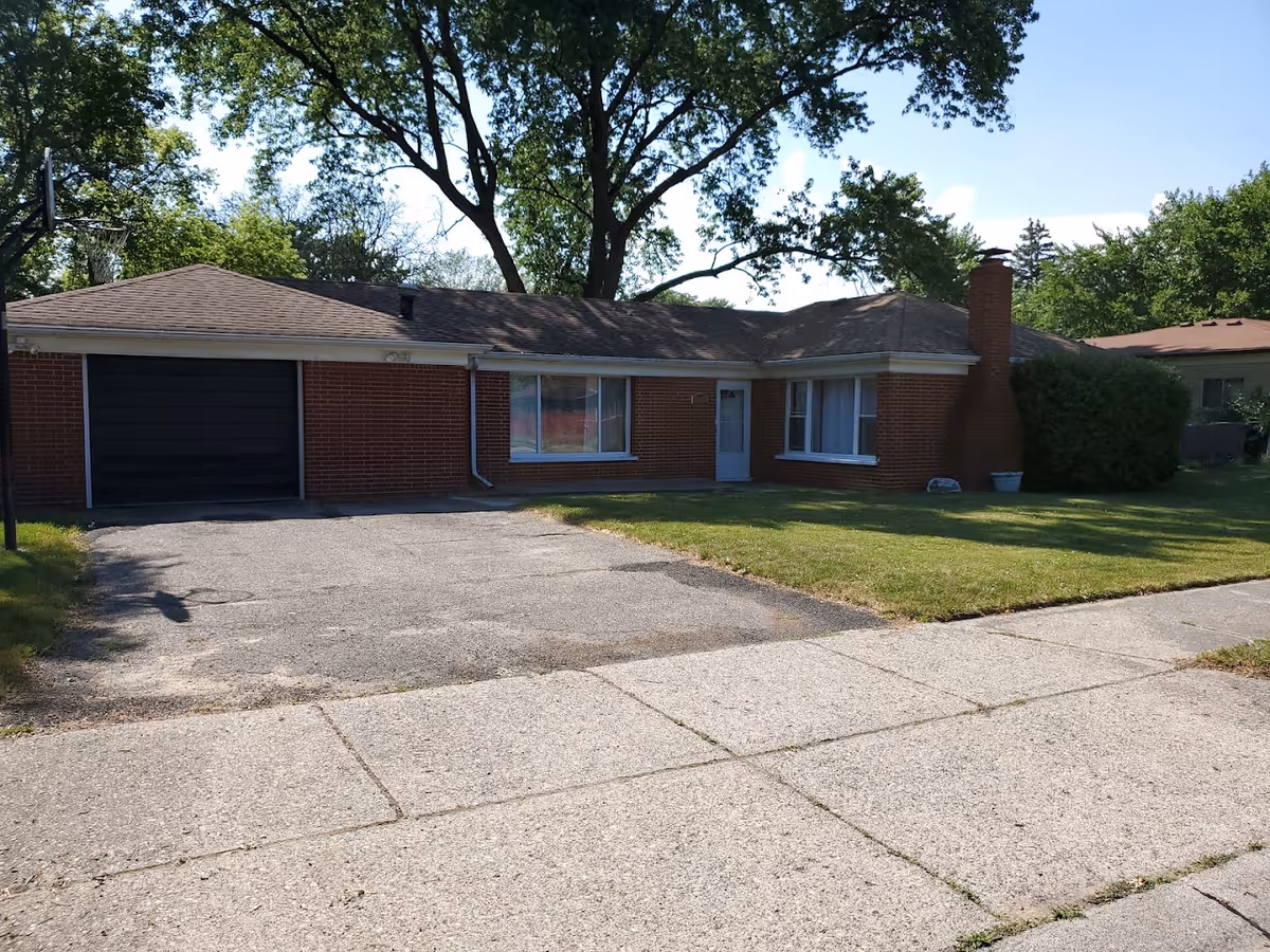Single-story brick house with a garage, large windows, a front door, a chimney, and a well-maintained lawn with trees in the background under a clear blue sky.