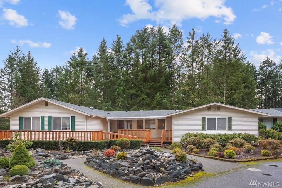 Exterior view of a single-story residential building with white siding and green shutters, surrounded by landscaped garden with rocks, shrubs, and trees under a blue sky with scattered clouds.