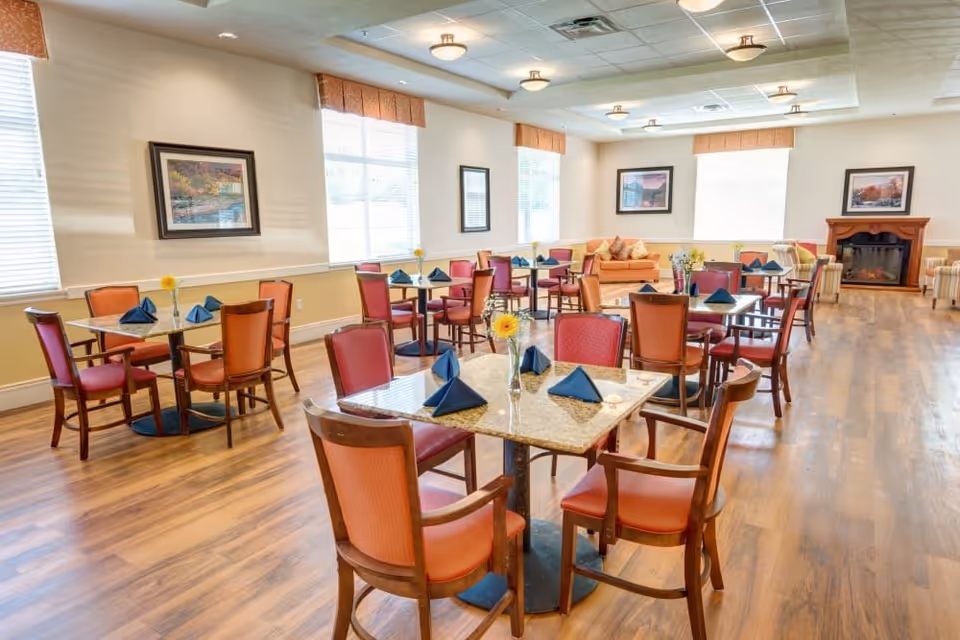 Bright dining room with multiple tables set with blue napkins, wooden chairs, and a seating area with sofas and a fireplace.
