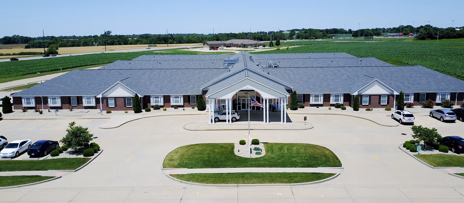 Aerial view of Garden Place Waterloo, a single-story senior living facility with a large covered entrance and an American flag in front. The building is surrounded by a paved parking lot with several cars parked, and green fields extend into the distance under a clear blue sky.