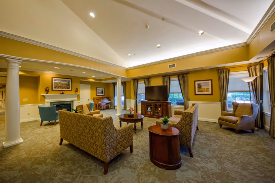 A spacious senior living facility common area with patterned sofas and armchairs arranged around a wooden coffee table and a TV on a wooden stand. The room features large windows with curtains, a fireplace with decorative items on the mantel, a piano in the corner, and warm yellow walls with white trim and columns.