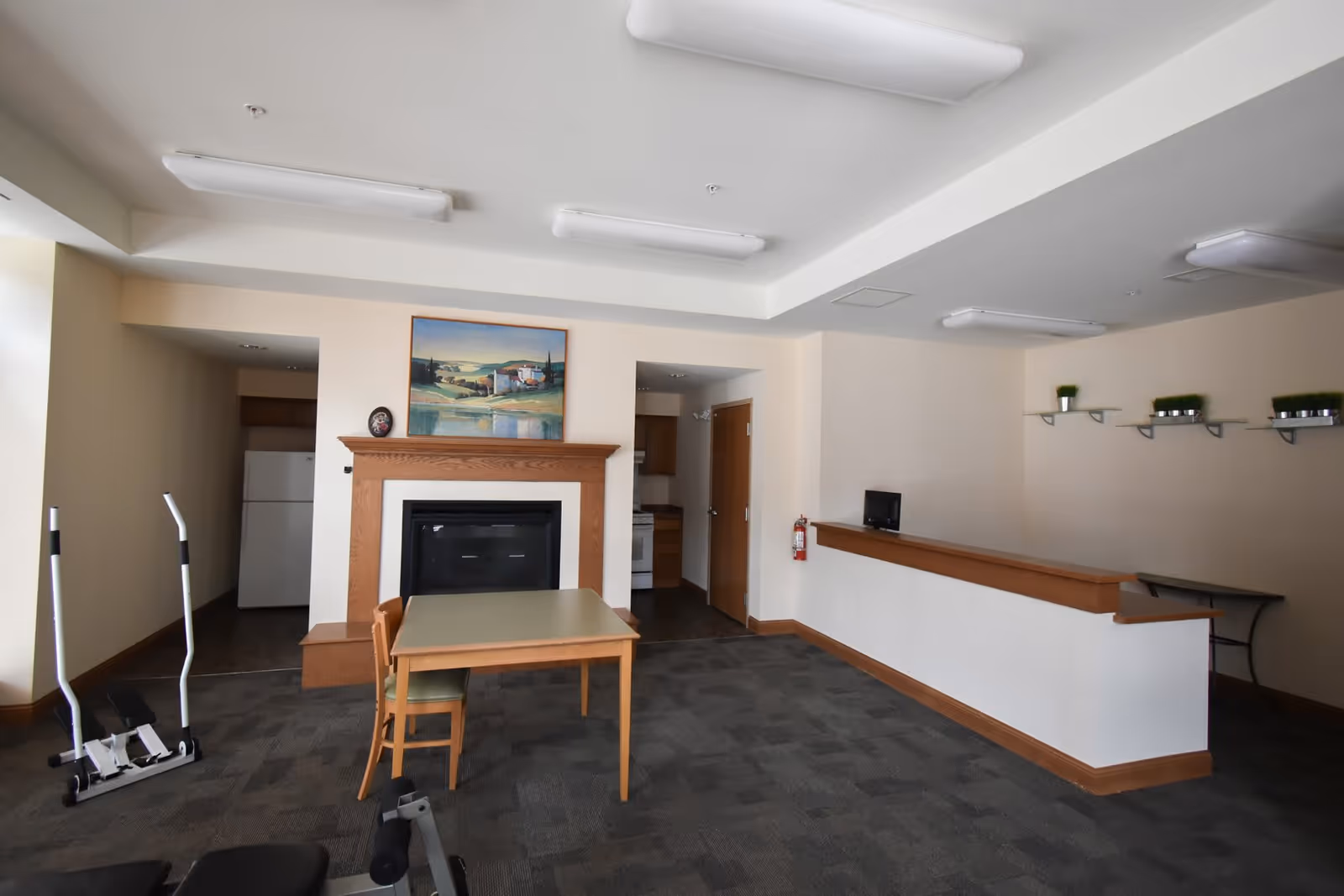 Interior room with a small table and two chairs in front of a fireplace with a landscape painting above it. To the left, there is exercise equipment and a refrigerator visible through an open doorway. On the right side, there is a counter with a small computer monitor and shelves with small potted plants on the wall above it.