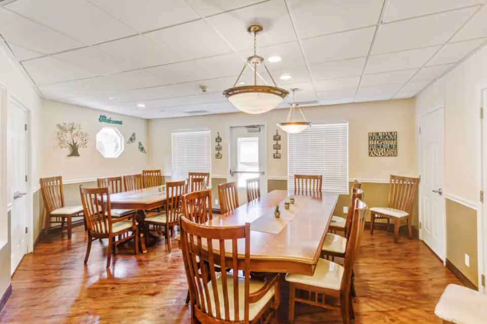 Bright communal dining room with long wooden tables, multiple wooden chairs, pendant lights, and wall decorations.
