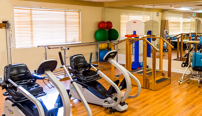 A rehabilitation exercise room with two recumbent exercise bikes, wooden handrails for walking support, colorful exercise balls stacked in the corner, a small basketball hoop, and mirrored walls reflecting the equipment and windows with blinds.