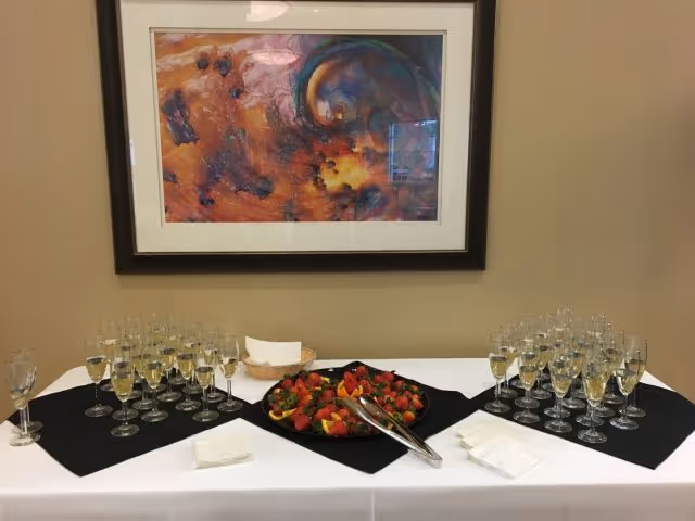 Table set with rows of champagne glasses and a platter of cut fruit beneath a framed abstract painting.
