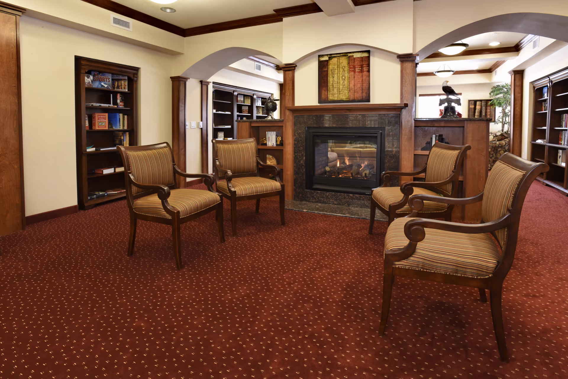 A cozy sitting area with four wooden armchairs upholstered in striped fabric arranged around a fireplace with a dark stone surround. The room features a red patterned carpet, cream-colored walls, wooden trim, built-in bookshelves filled with books, and decorative items. The ceiling has recessed lighting and wooden beams.