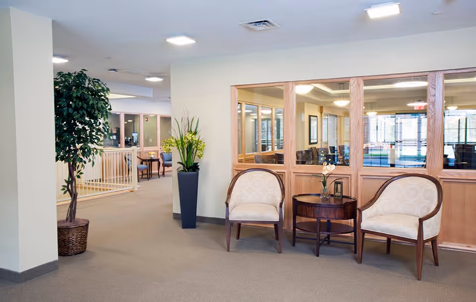 A seating area in a senior living facility with two beige upholstered chairs and a small round wooden table between them, decorated with a small vase of flowers. There are large windows with wooden frames behind the chairs, and a tall potted plant and a tall vase with flowers are placed nearby. The space is well-lit with ceiling lights and has beige carpeting.