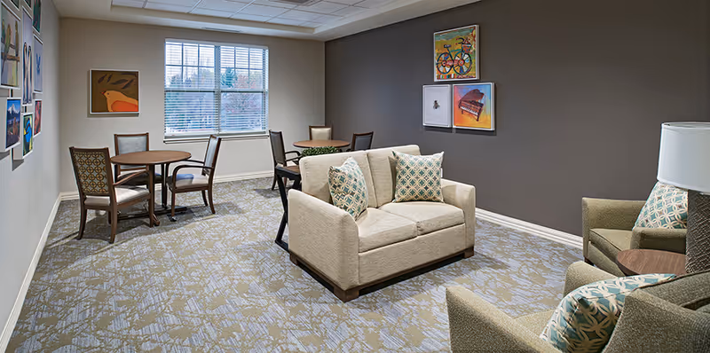 A cozy common area in a senior living facility featuring a beige loveseat with patterned pillows, two armchairs with matching pillows, and two small round tables with chairs near a window. The walls are decorated with colorful framed artwork, and the room has a patterned carpet and neutral wall colors.