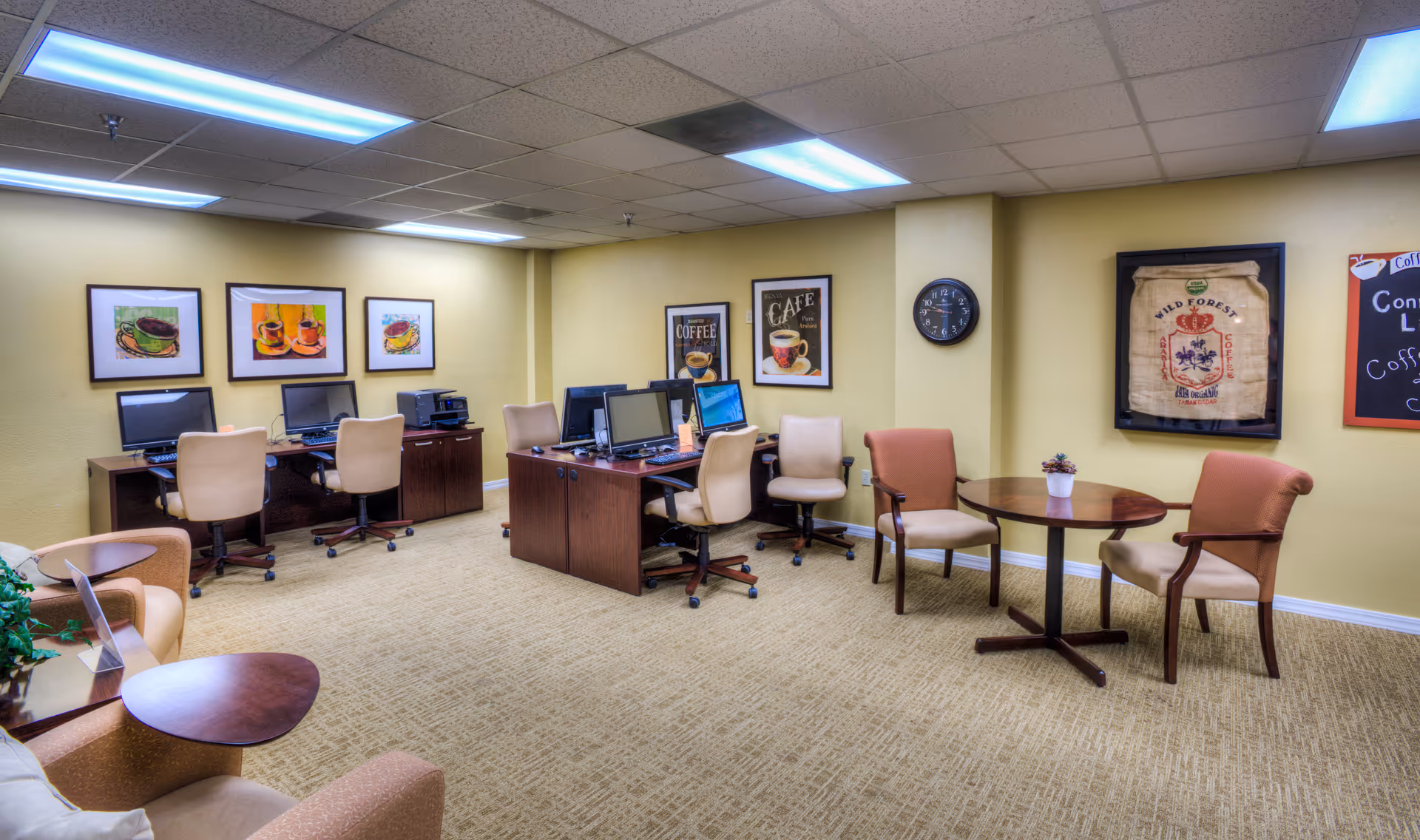 A well-lit interior common area with computer desks and chairs, round tables with seating, and coffee-themed wall art.
