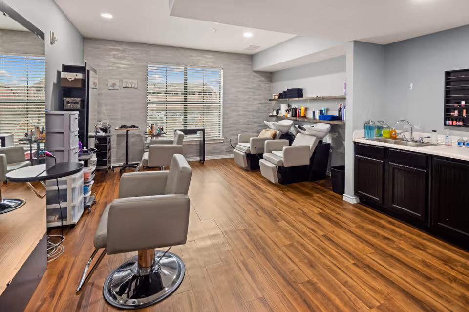 Interior view of a salon area in a senior living facility with wooden flooring, salon chairs, hair washing stations, a large mirror, and shelves with hair care products and towels. A window with blinds lets in natural light.