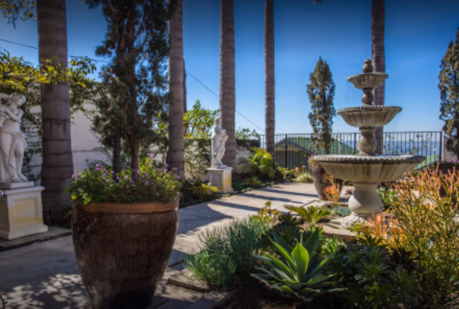 Outdoor garden area with tall palm trees, a large tiered stone fountain, various green plants and shrubs, and classical statues on pedestals under a clear blue sky.