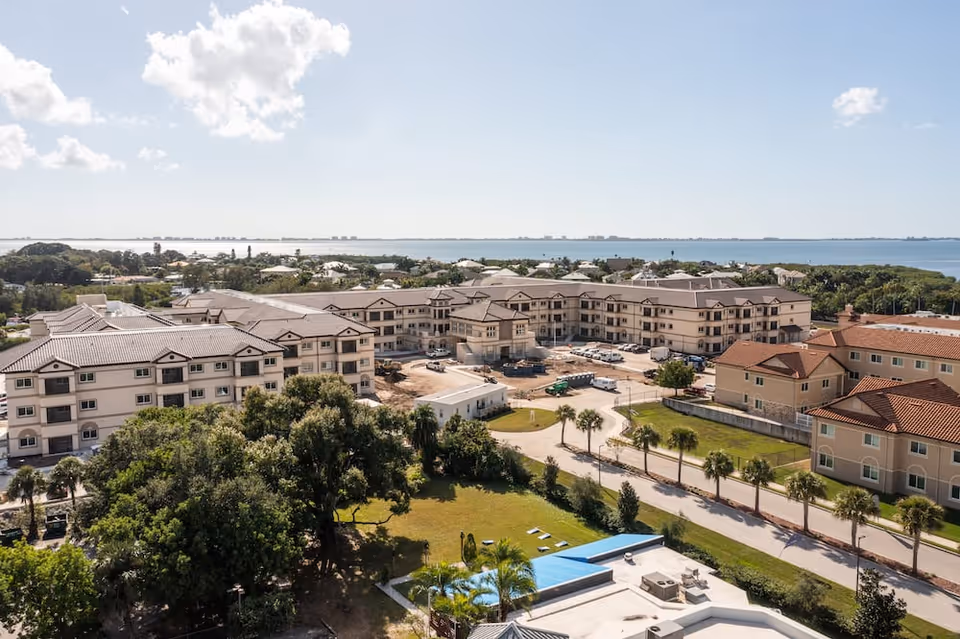 Aerial view of Discovery Village At Sarasota Bay senior living facility with multiple connected buildings, surrounded by trees and greenery, with a body of water visible in the background under a partly cloudy sky.