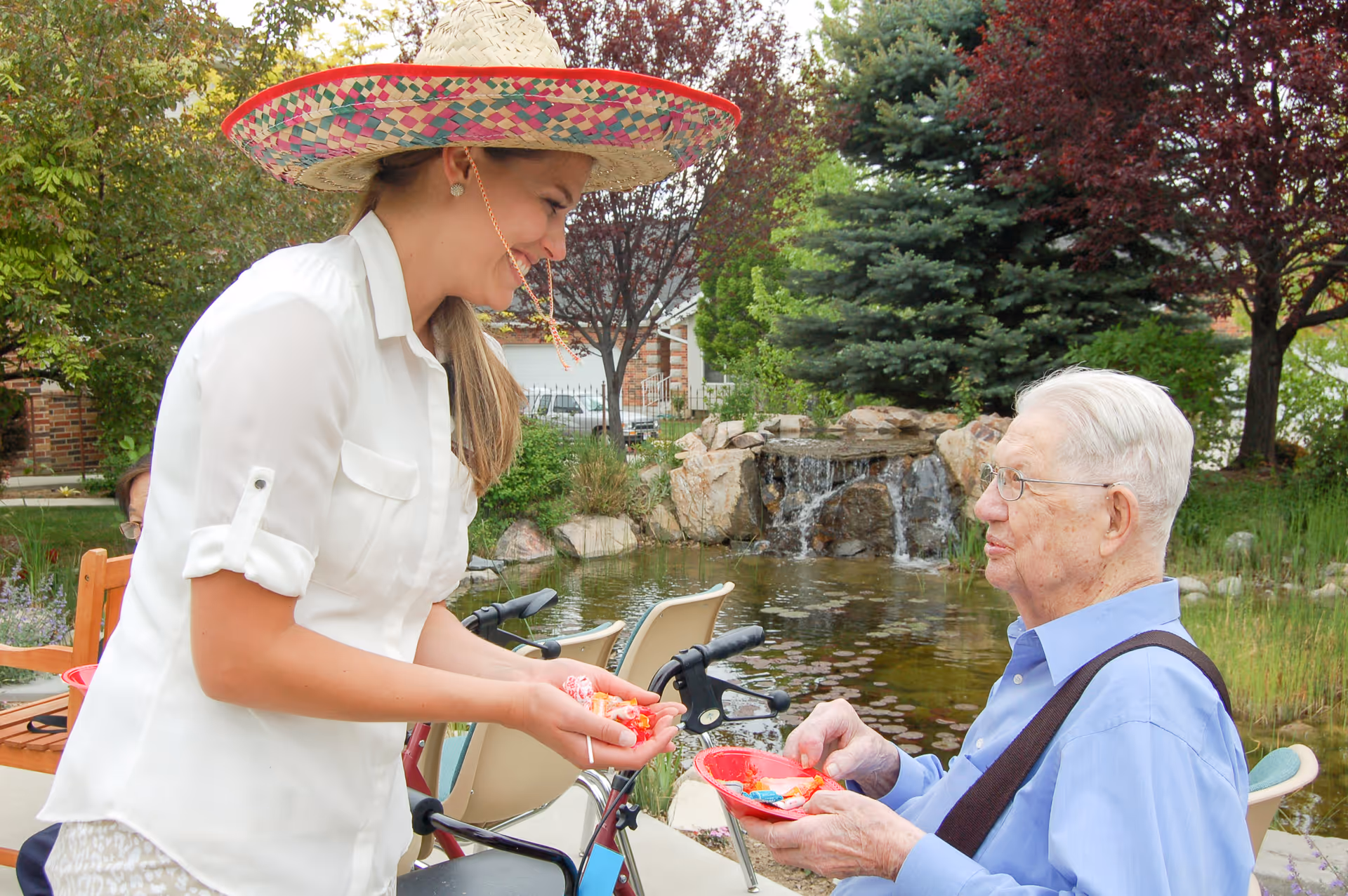 A young woman wearing a colorful sombrero and a white shirt is smiling and handing out candy to an elderly man with glasses and a blue shirt sitting outdoors near a pond with a small waterfall and surrounded by trees and greenery.